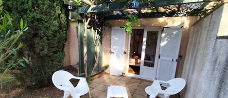 Un patio con sillas de plástico blanco y una mesa pequeña, una pérgola de madera, y una puerta que da a una habitación.