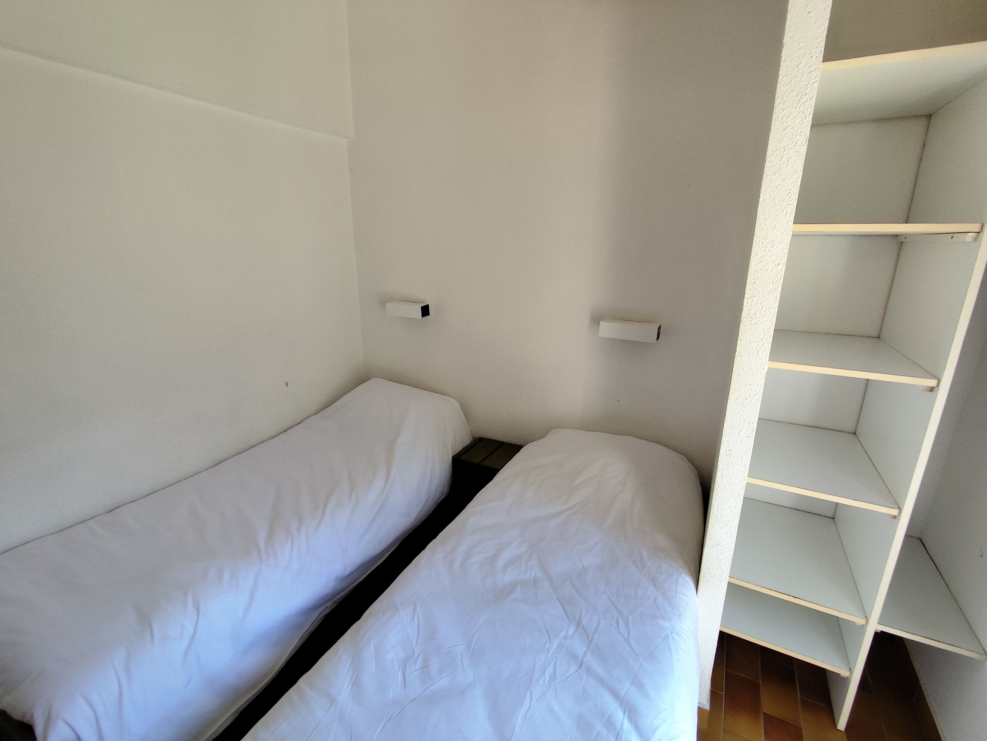 A small, single-room with two single beds, a white wall, and a built-in shelving unit.