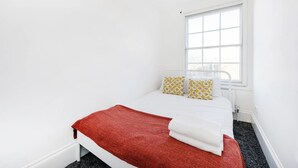 Une petite chambre blanche, avec un lit, une fenêtre et un radiateur.