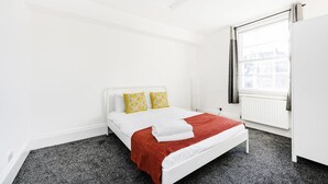 Un lit blanc avec une couverture rouge, deux oreillers jaunes et une fenêtre donnant sur des immeubles.