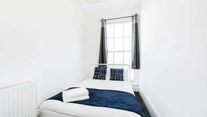 Une petite chambre blanche, avec un lit simple, une fenêtre, un radiateur blanc et un luminaire au plafond.