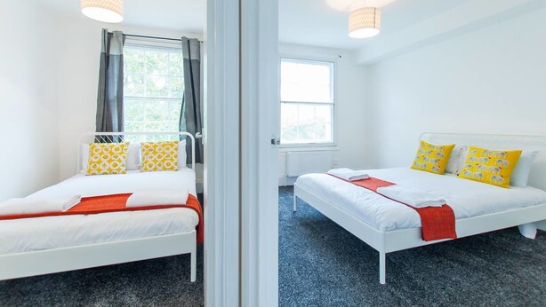 Une chambre double avec deux lits, chacun doté de draps blancs et d’oreillers jaunes, une fenêtre avec des rideaux et un luminaire au plafond.