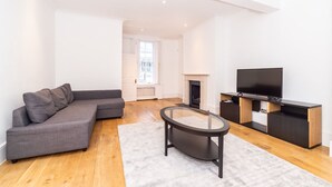 A modern living room with a grey sectional sofa, a glass coffee table, a flat-screen TV, and a fireplace. - PML Apartments Notting Hill (London)