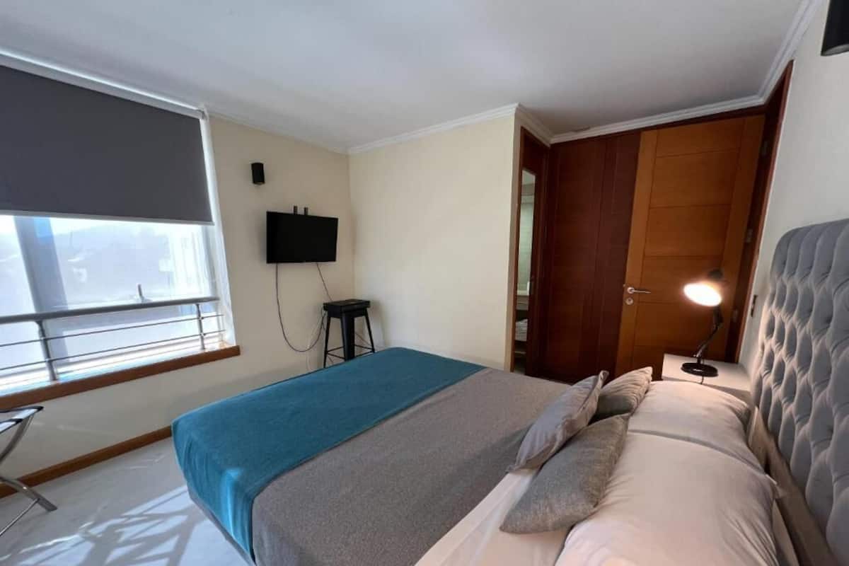 A bedroom with a bed, a TV, a wooden wardrobe, and a window with blinds.