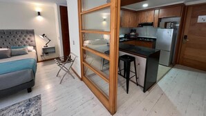 Un espace de vie compact comprenant un coin cuisine, un lit avec une tête de lit capitonnée et un coin repas avec un tabouret de bar.