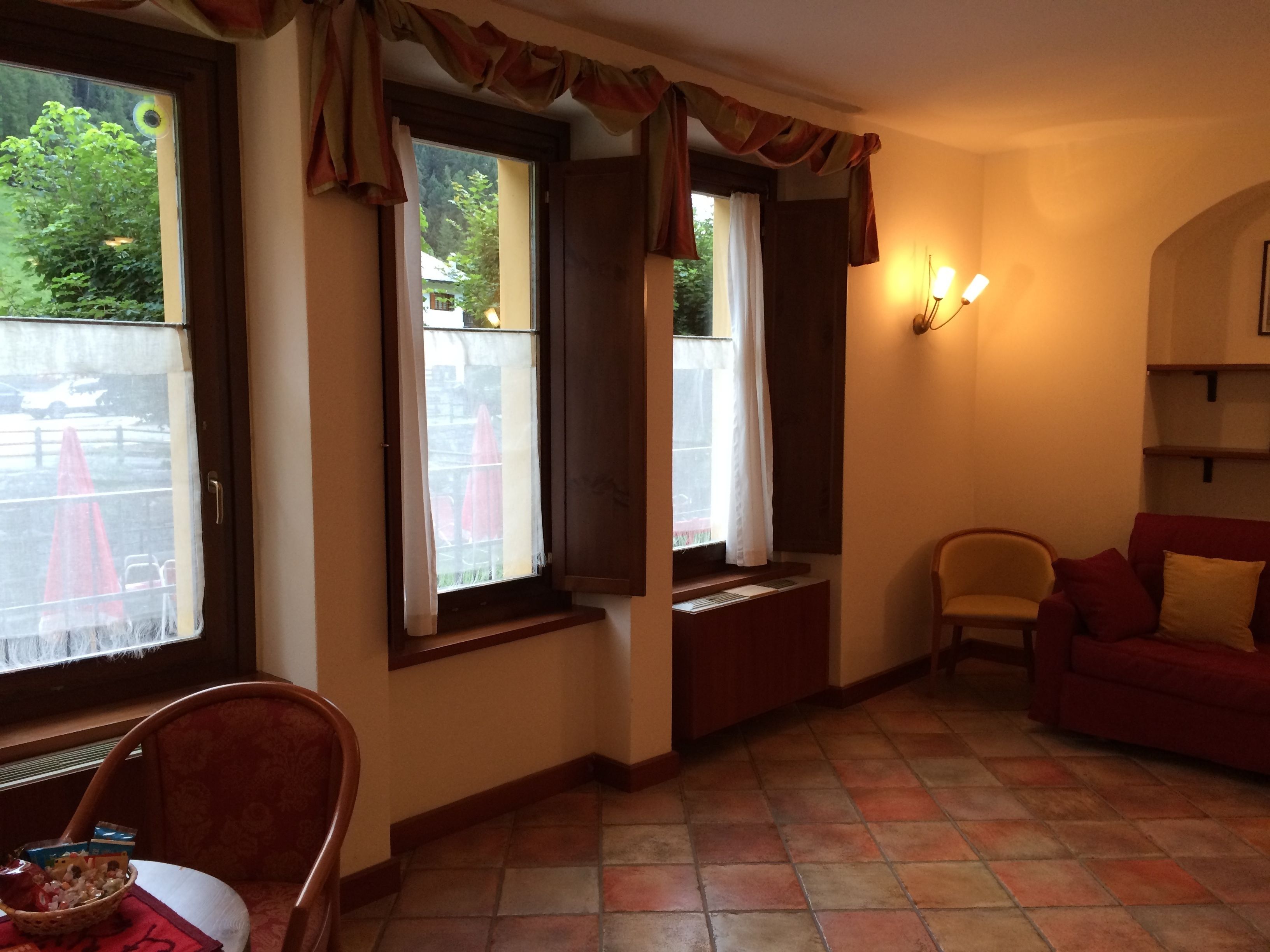 A cozy living room with a red sofa, a wooden chair, and a dining table with a basket. There are windows with curtains and a wall-mounted light fixture.