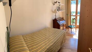 A bedroom with a bed, a desk with a lamp, a chair, and a piano.