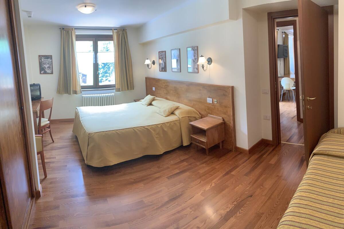 A hotel room with a wooden floor, a bed with a beige bedspread, a nightstand, a desk with a chair, and a window with curtains.