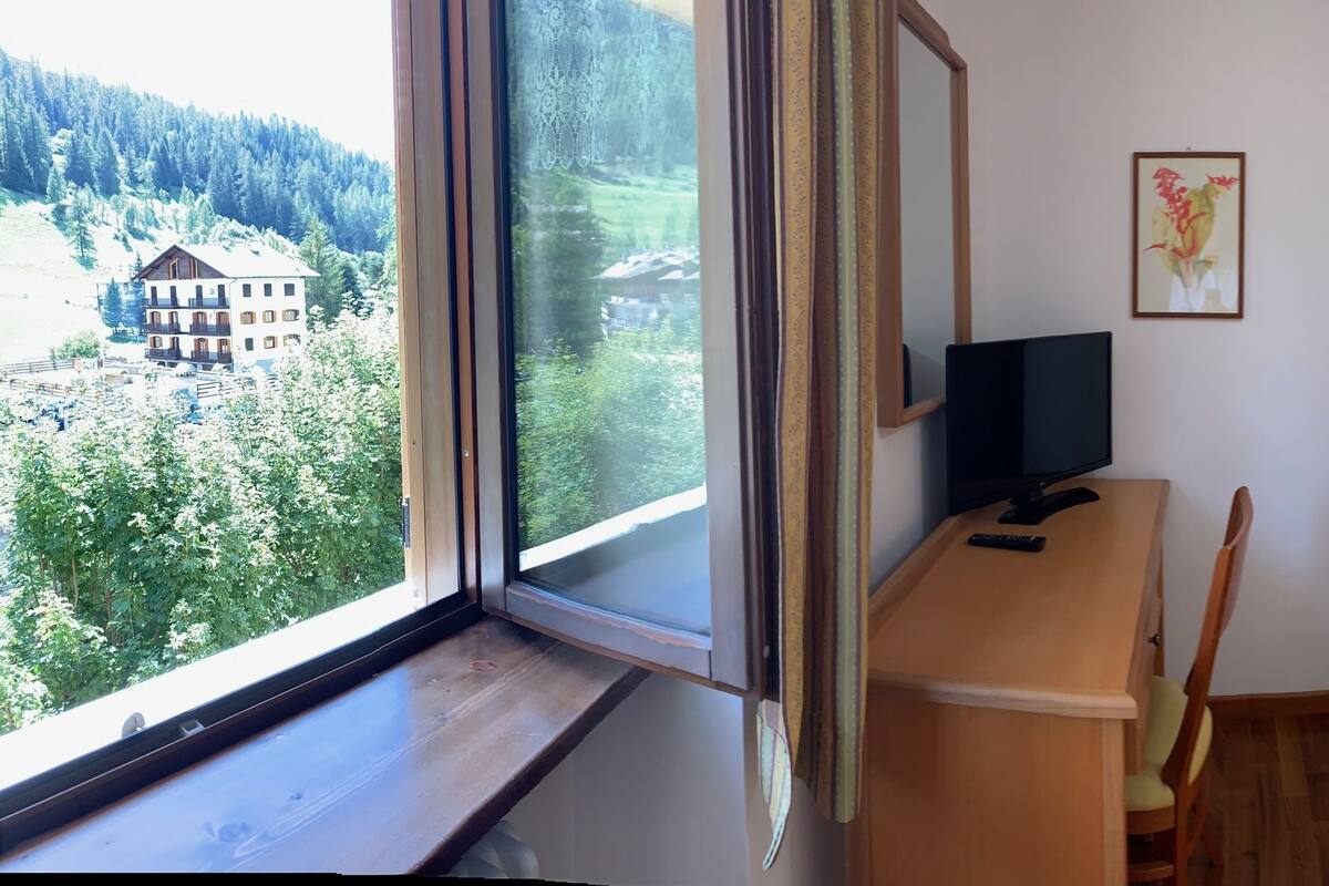 A hotel room with a bed, a desk with a chair, a television, and a wardrobe. There is a window with a view of a mountain and a building.