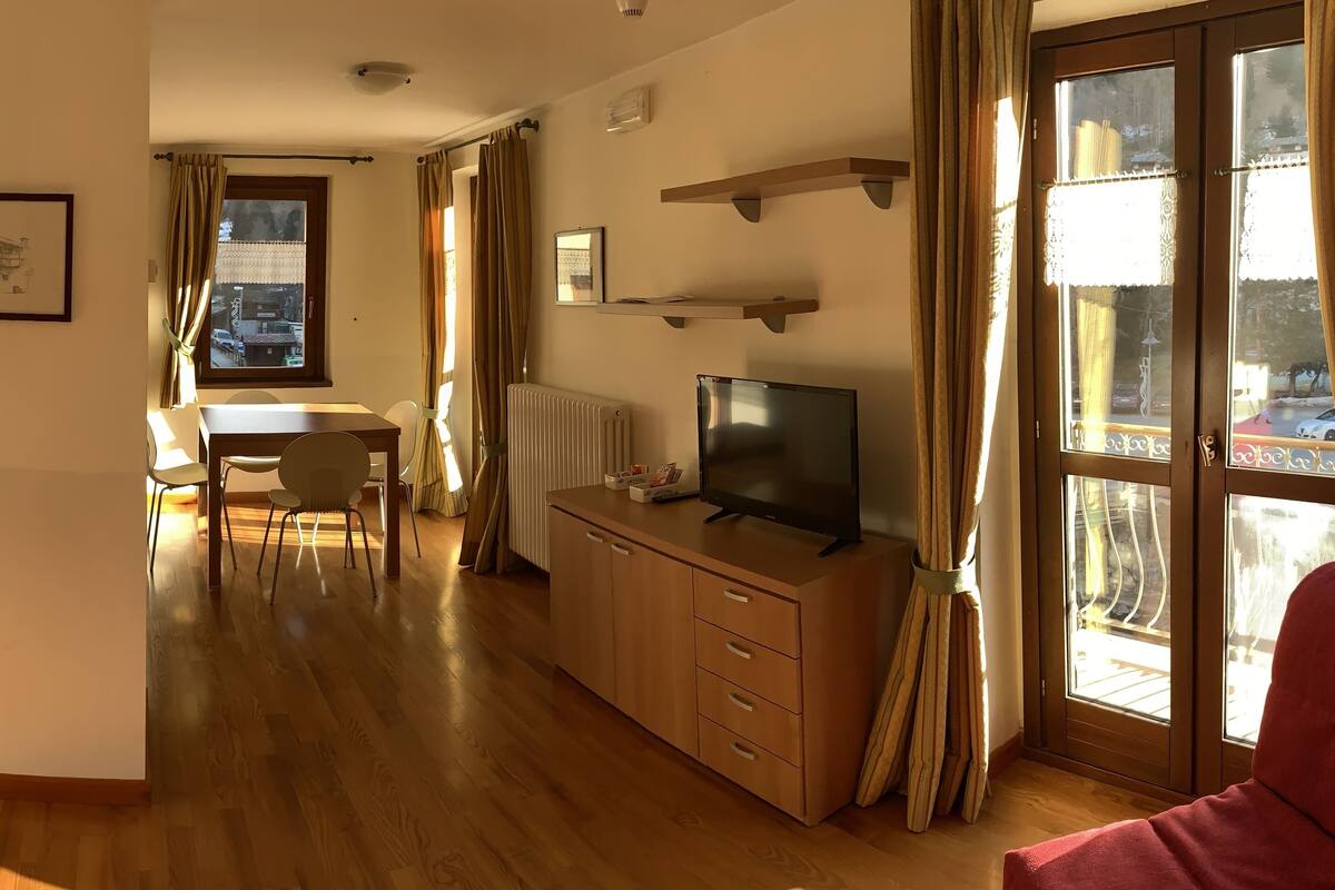 A modern living room with a wooden floor, a TV on a wooden cabinet, a dining area with a table and chairs, and a large window with curtains.
