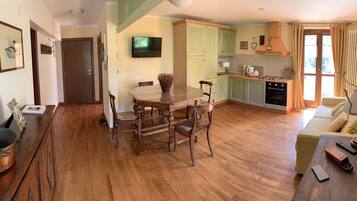 A spacious living area with a wooden dining table, chairs, and a sofa. A kitchen with green cabinets and a stove is visible in the background.