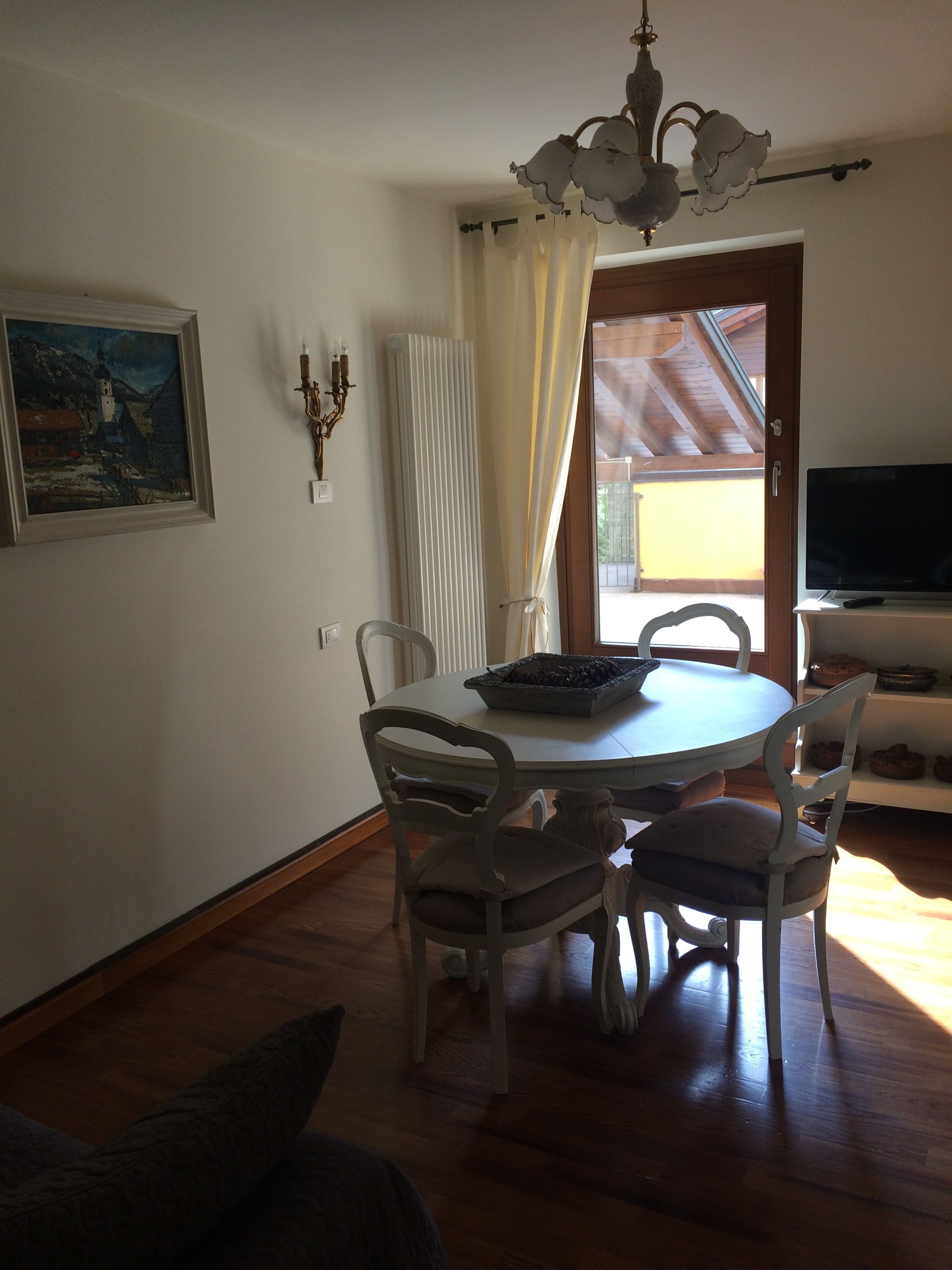 Ein Essbereich mit einem runden Tisch, weißen Stühlen, einer Kronleuchter, einem Gemälde an der Wand und einem Fernseher in der Ecke.