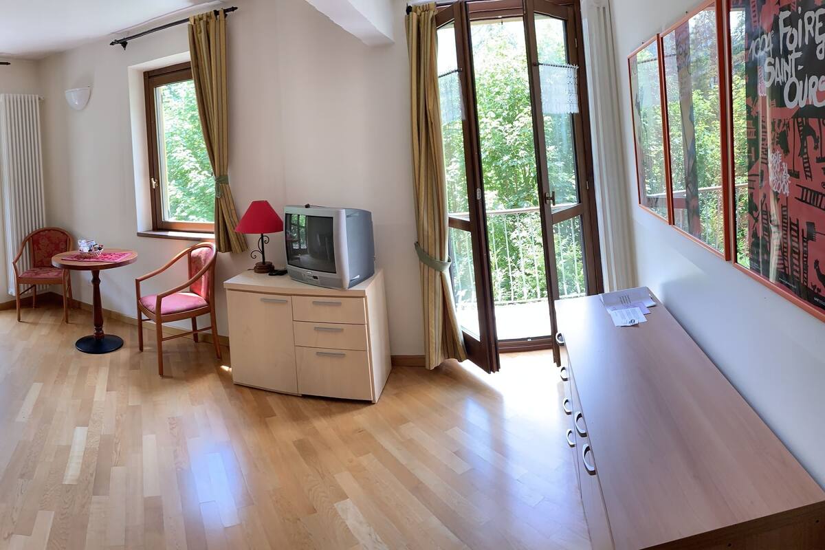 A cozy living room with a red sofa, a dining table with chairs, a small round table with a chair, a TV on a cabinet, and large windows with curtains.