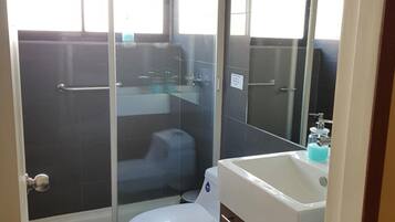 Un baño compacto con cabina de ducha de vidrio, inodoro blanco y lavamanos con dispensador de jabón.