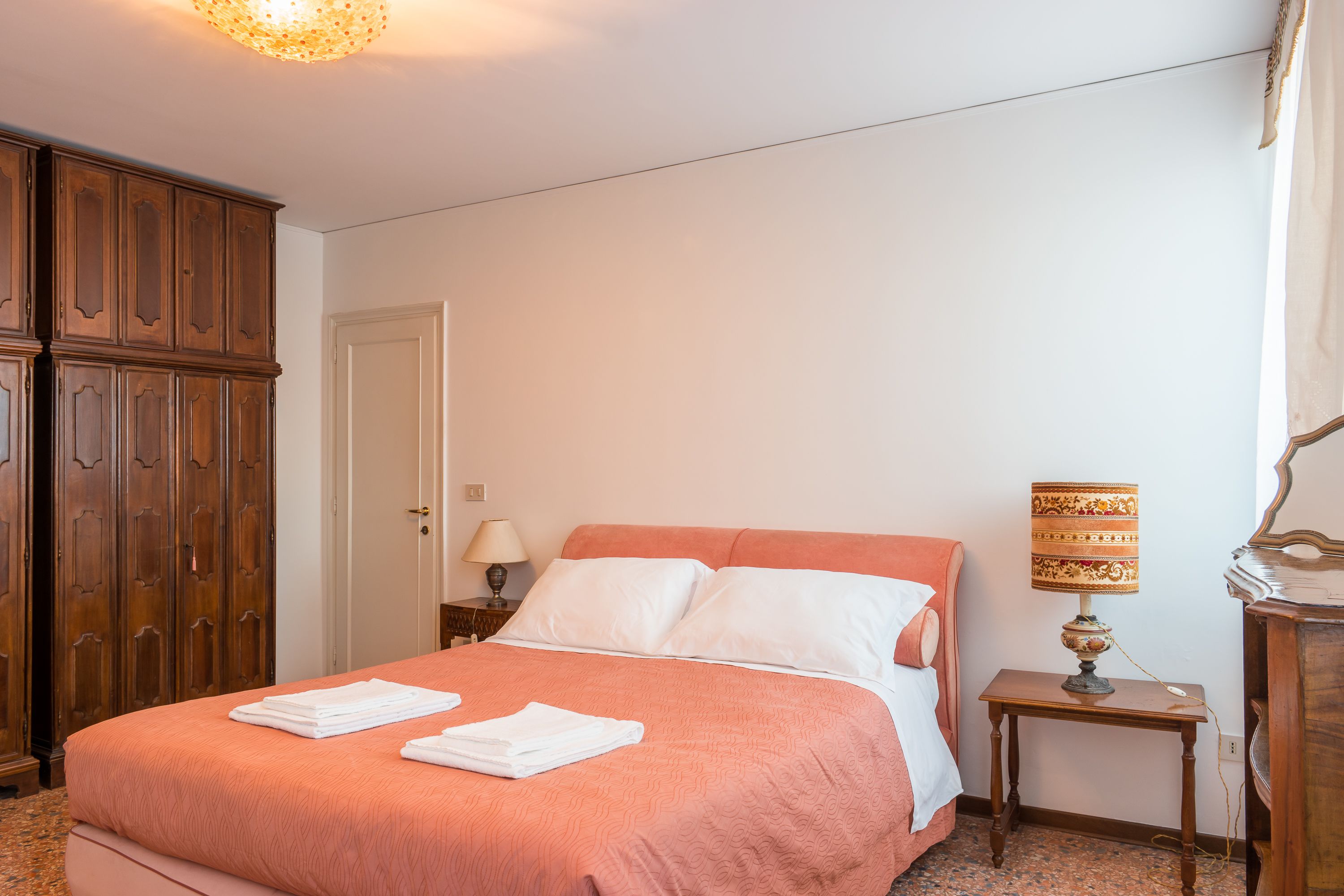 A neatly made bed with white linens and a pink bedspread, a wooden wardrobe, a bedside table with a lamp, and a door in the background.