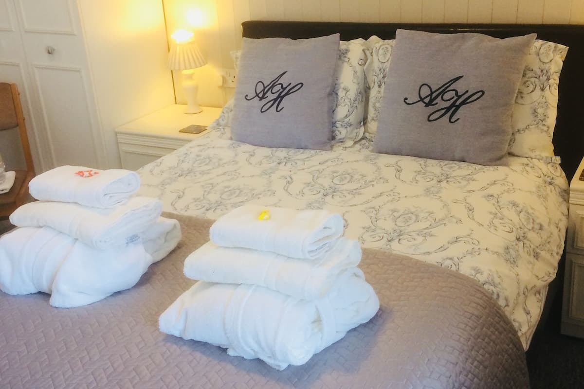 A neatly made bed with folded towels and two pillows with embroidered text, a bedside lamp, and a white wardrobe.