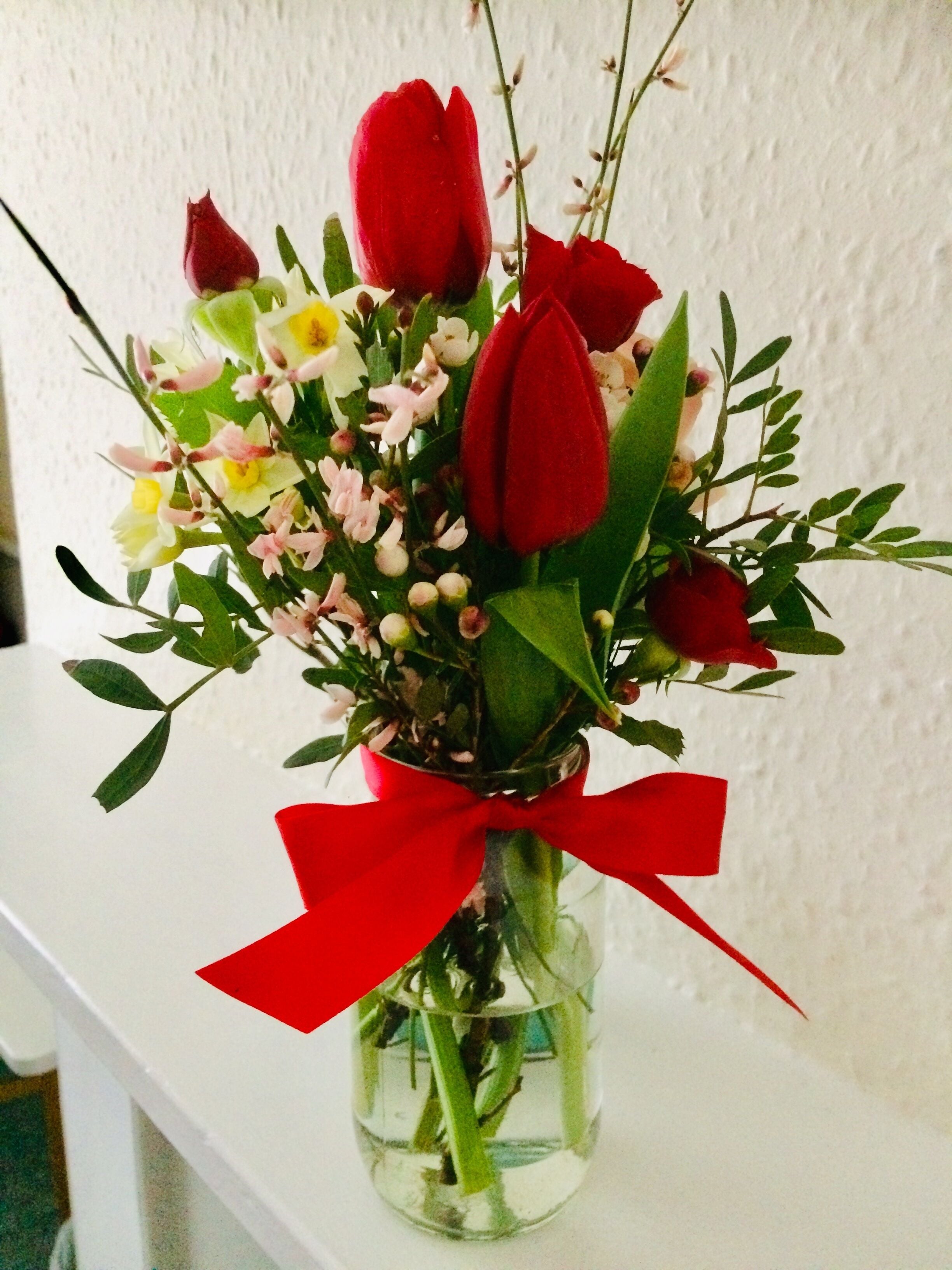 A clear glass vase with a red ribbon, containing red tulips, white flowers, and green leaves.