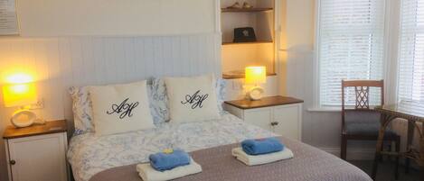 A neatly arranged bedroom with a bed, two bedside lamps, a nightstand, a chair, a small table, a shelf with decorative items, and a framed picture on the wall.