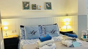 Una cama bien hecha con colcha acolchada, dos almohadas con texto bordado, una mesita de noche con lámpara y una estantería con fotografías enmarcadas.