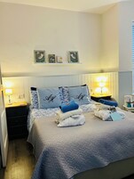 Cama bem arrumada com edredom acolchoado, duas almofadas com texto bordado, mesa de cabeceira com abajur e prateleira com quadros.