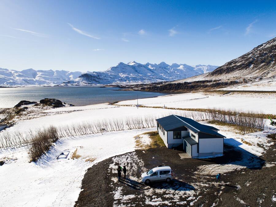 A small, single-story building with a sloped roof, situated on a snow-covered landscape with a body of water and mountains in the background.