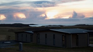 Un edificio moderno con techo plano y varias ventanas, ubicado en un área verde con vista al mar y nubes en el cielo.