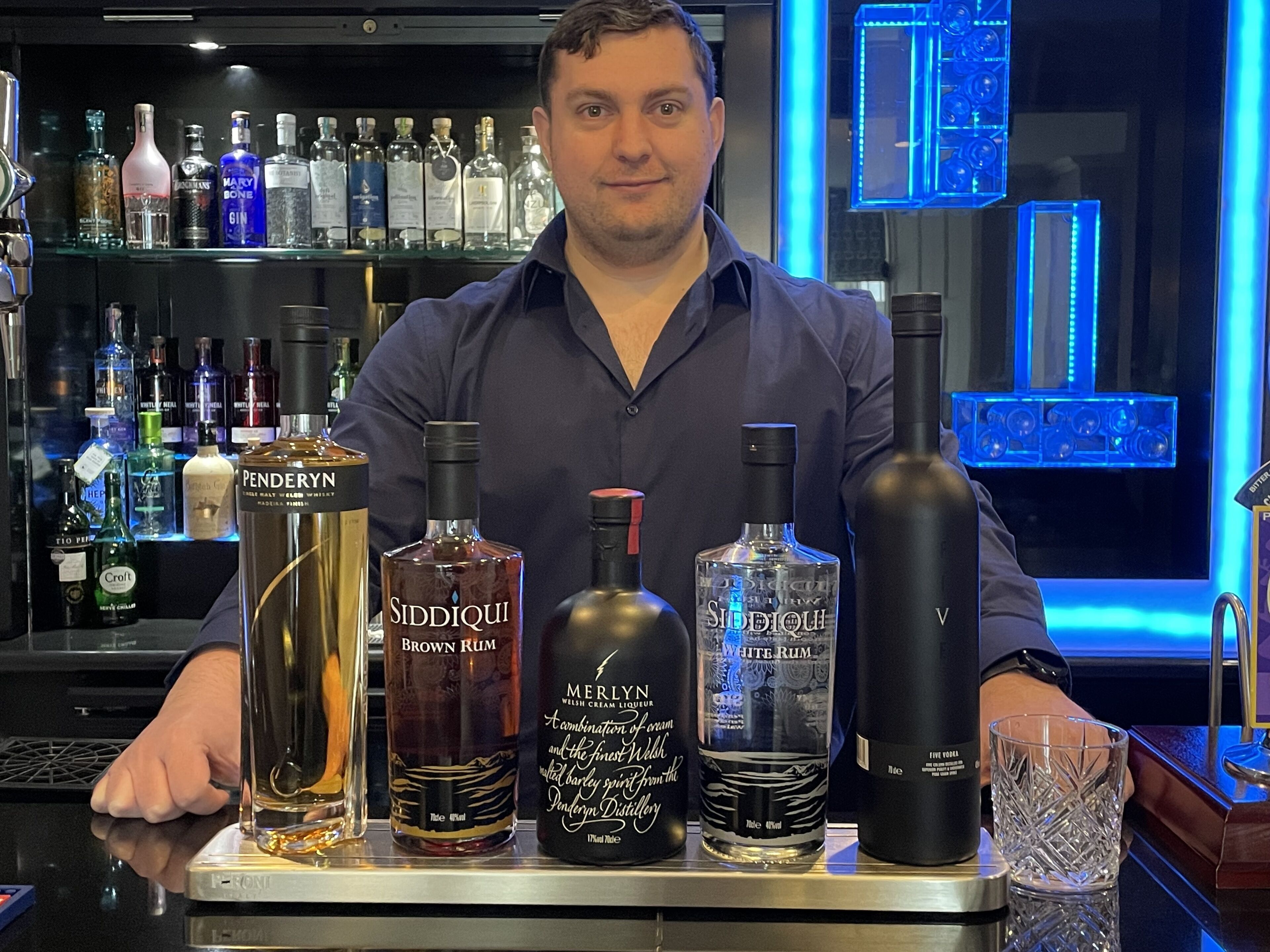 A bartender standing behind a bar with various bottles of alcohol, including rum and gin.