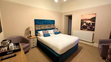 A hotel room with a large bed, a bedside table with a lamp, a chair, a desk with a tray of toiletries, and a painting on the wall.