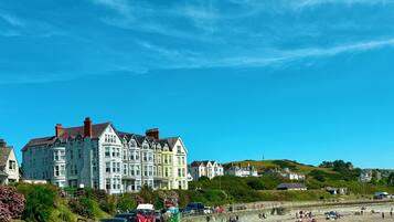A coastal town with a row of colorful Victorian-style buildings, a sandy beach with people, and parked cars along the promenade.