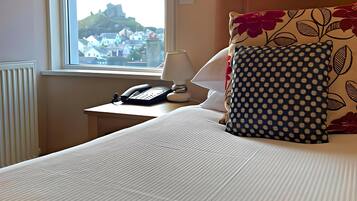 A hotel room with a bed, a nightstand with a lamp and phone, a window with a view of houses, and a radiator.