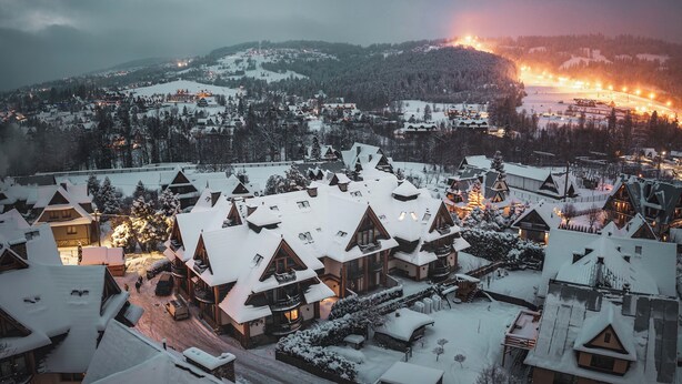 Un pueblo cubierto de nieve con edificios de estilo chalete tradicional, una pista de esquí al fondo y un brillo cálido de las luces.