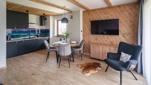 Et moderne køkken med mørke skabe, et spiseområde med et rundt bord og stole, et stort fjernsyn monteret på en trævæg, og en hund, der ligger på gulvet.