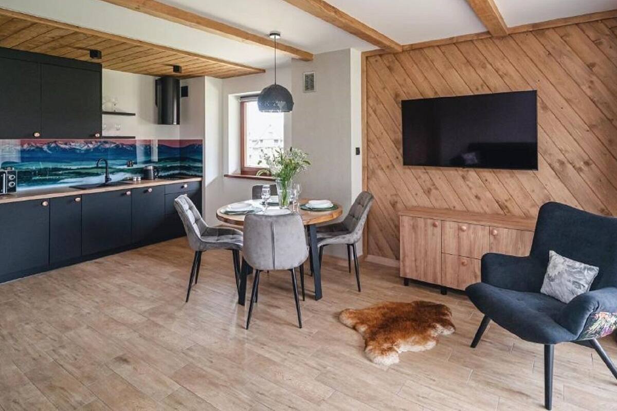 Une cuisine moderne avec des meubles de couleur sombre, un coin repas avec une table ronde et des chaises, un grand téléviseur fixé à un mur en bois, et un chien allongé sur le sol.