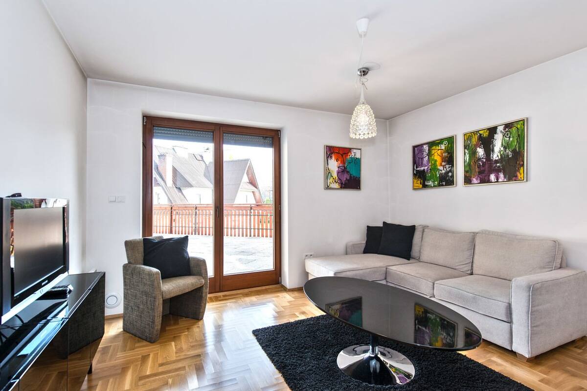 Un salon moderne avec un canapé, un fauteuil et une table basse. On y trouve une télévision et un haut-parleur sur le côté gauche. La pièce est dotée d’une grande porte-fenêtre coulissante qui donne sur un espace extérieur.