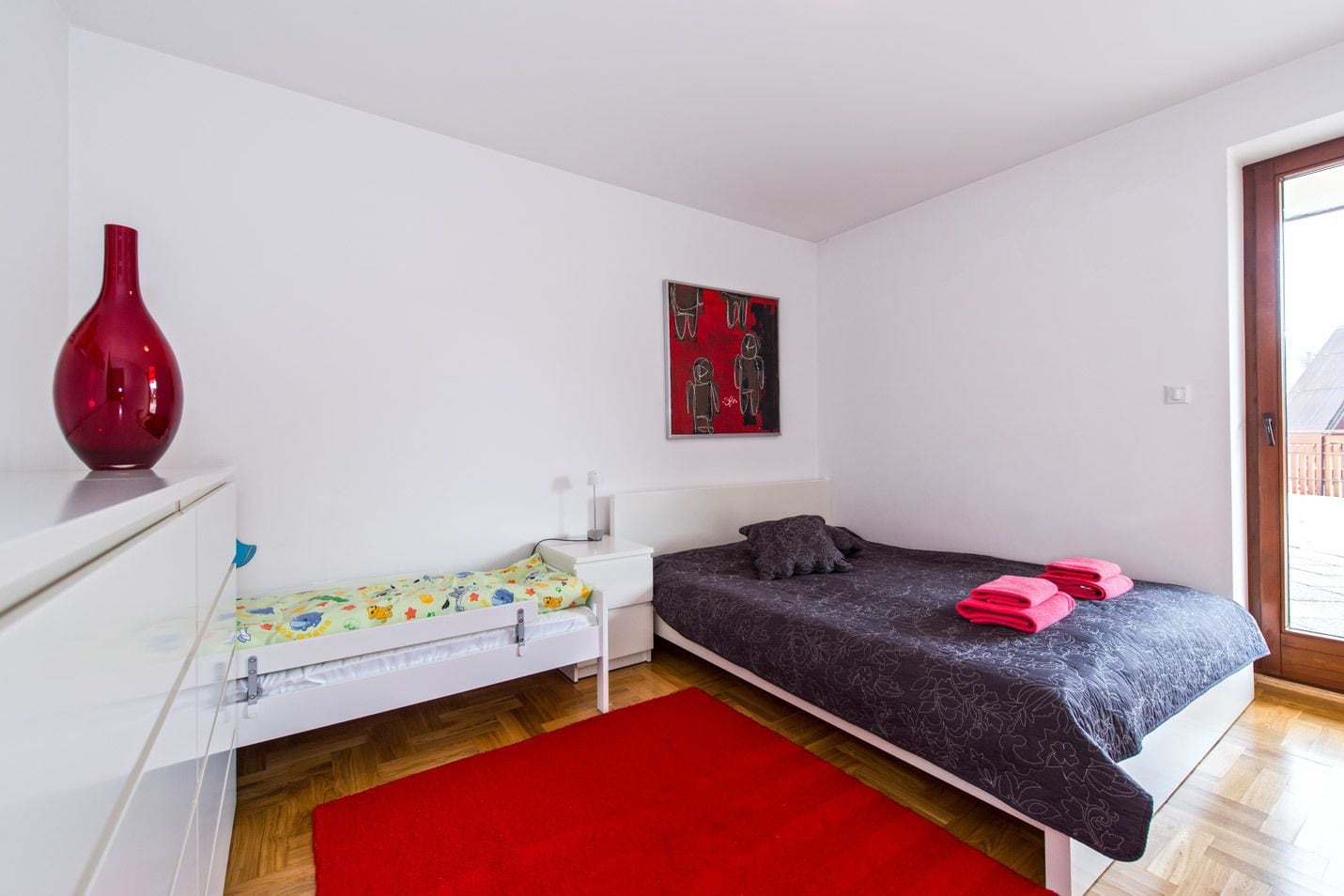 Un dormitorio con una cama individual, una mesita de noche, un jarrón rojo y un cuadro en la pared.