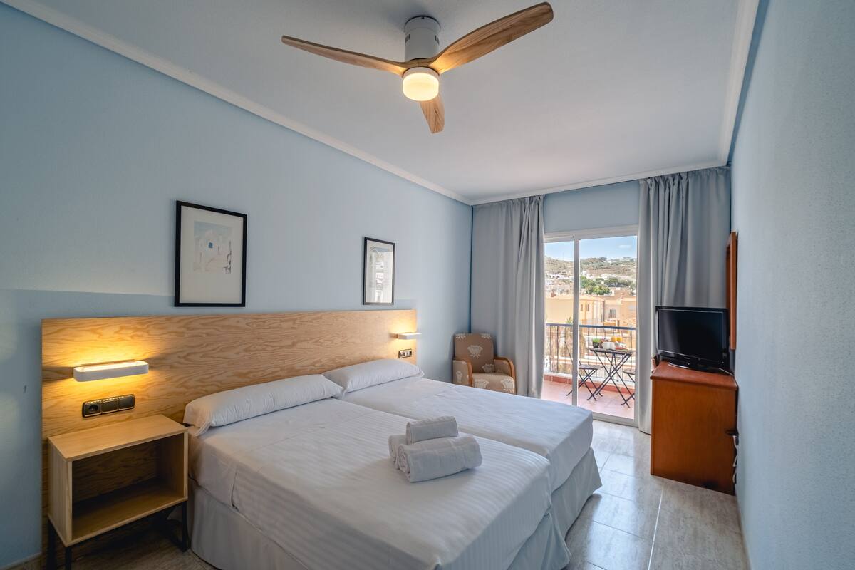Une chambre d’hôtel avec un grand lit, un ventilateur de plafond, une télévision et un balcon offrant une vue sur la ville.