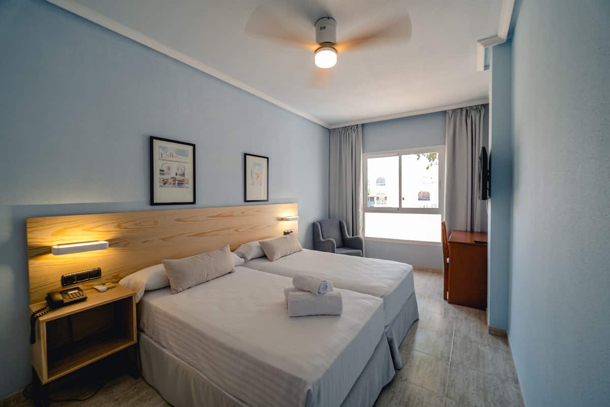 Une chambre d’hôtel avec deux lits, une tête de lit en bois, une table de chevet avec un téléphone, un bureau et une chaise.