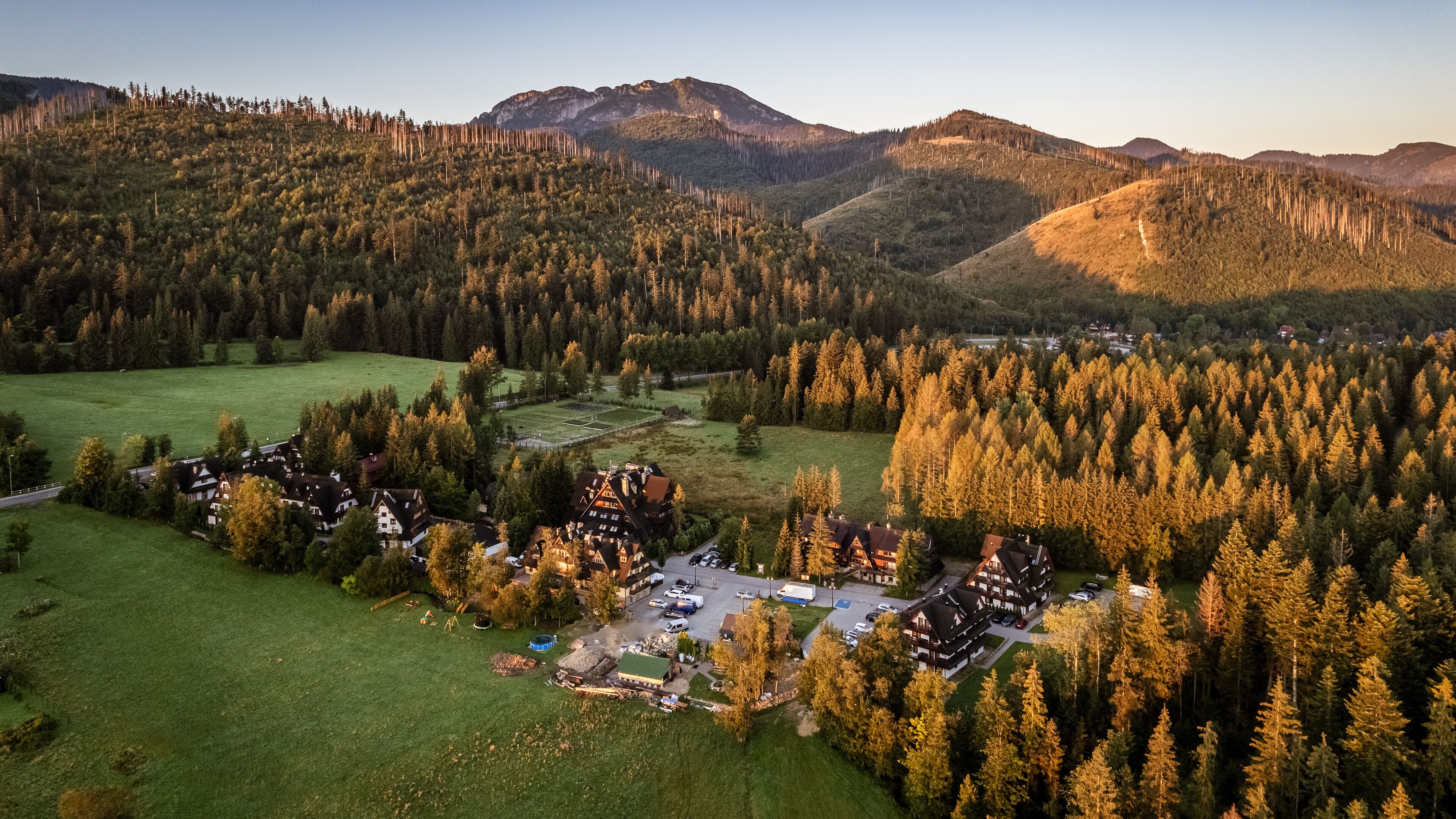An aerial view of a resort with multiple buildings, a parking area, and a forested landscape with mountains in the background.