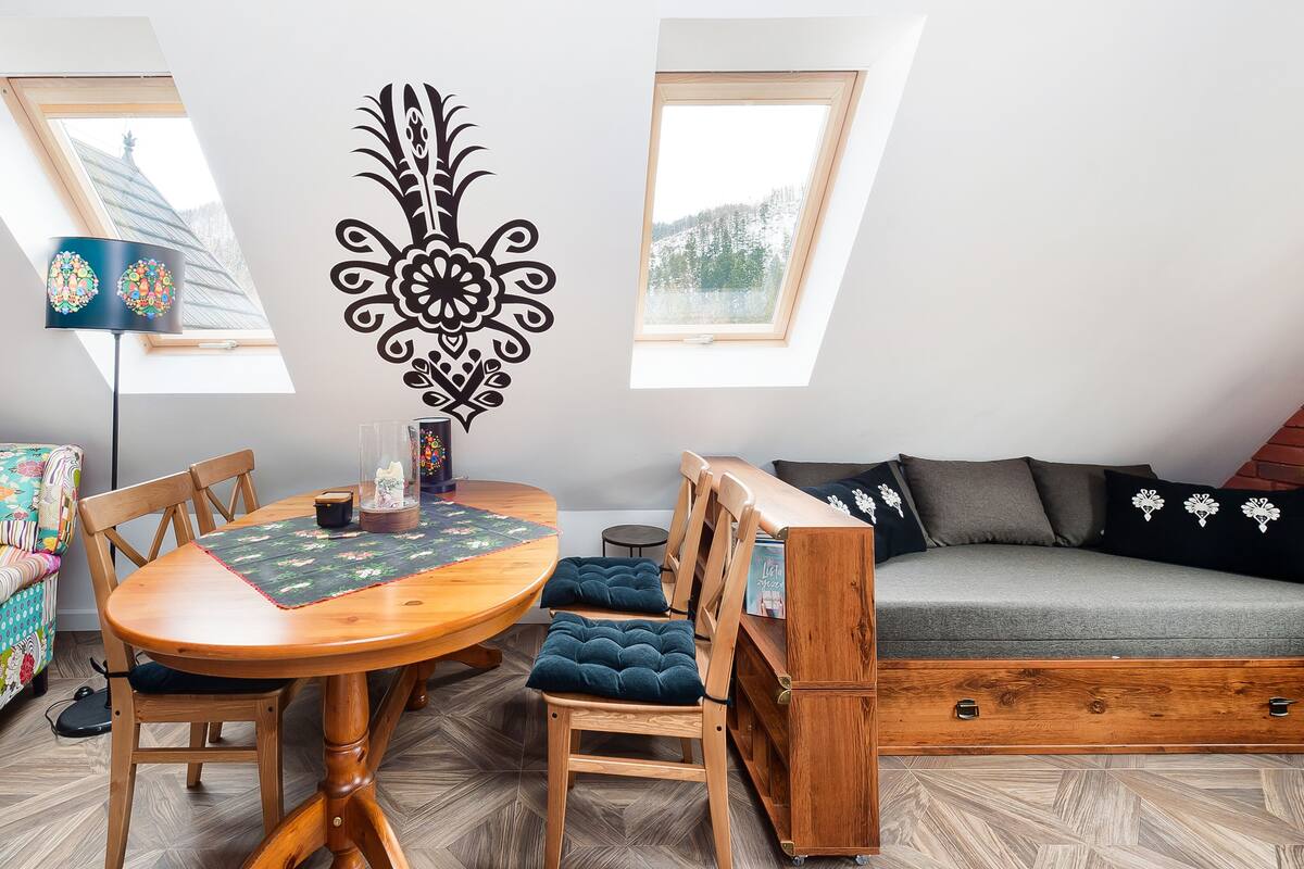 Een gezellige loft-achtige woonruimte met een houten eettafel, een kleurrijke bank en een grijze zithoek.