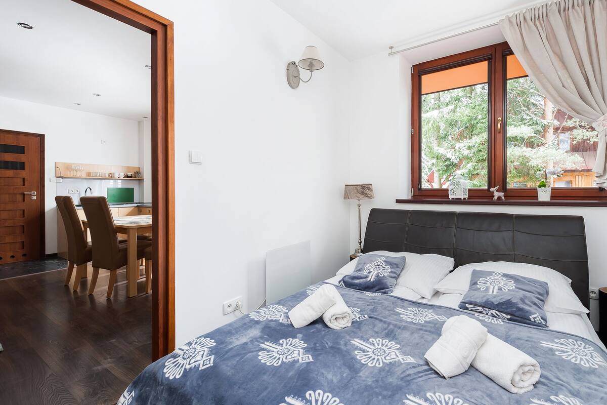 Een slaapkamer met een bed, een raam met gordijnen, een nachtkastje en uitzicht op een keuken en eethoek.