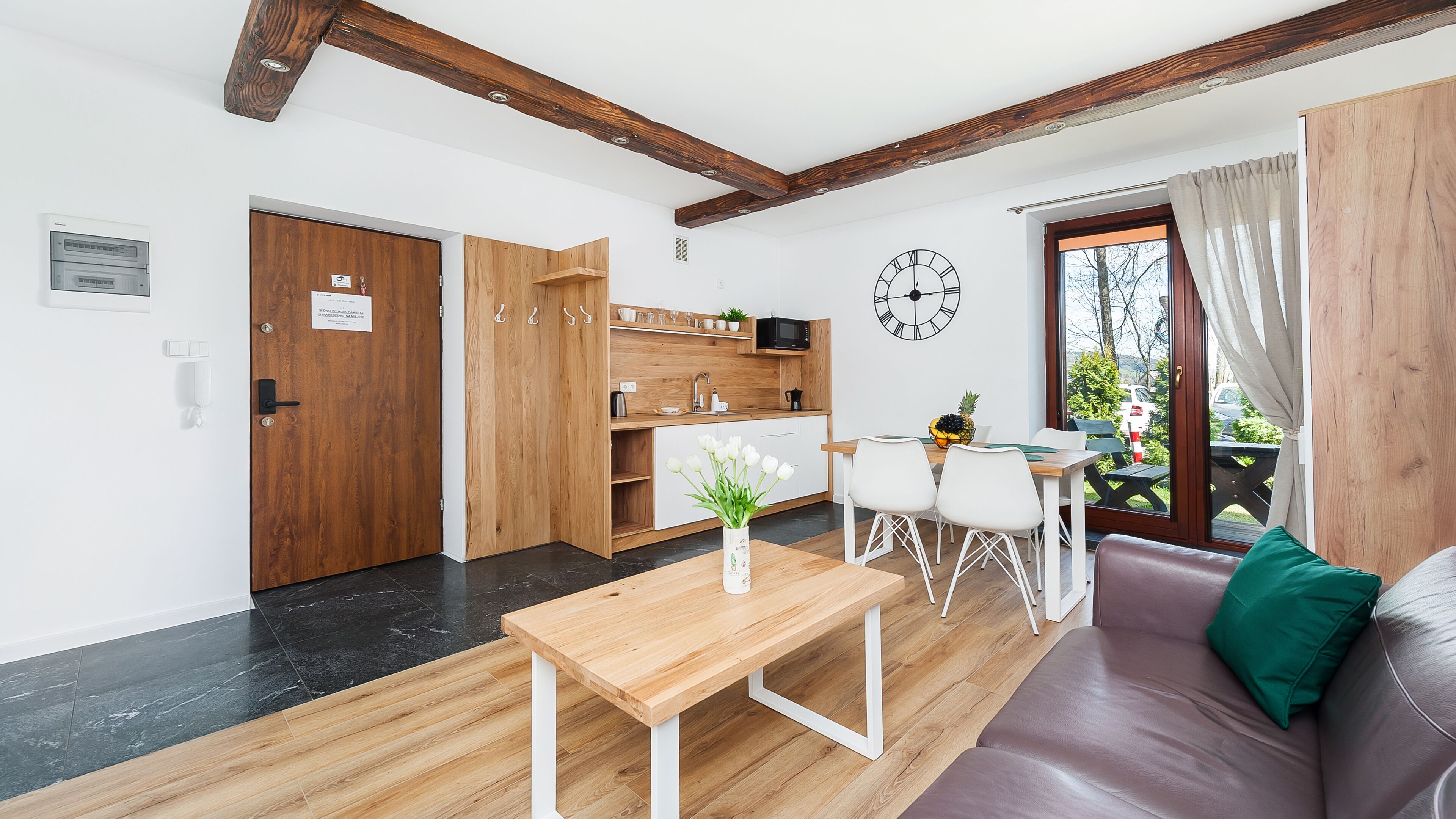 Un soggiorno moderno con pavimento in legno, un divano, un tavolo da pranzo con sedie, angolo cottura con mobili in legno e un'ampia finestra con tende.