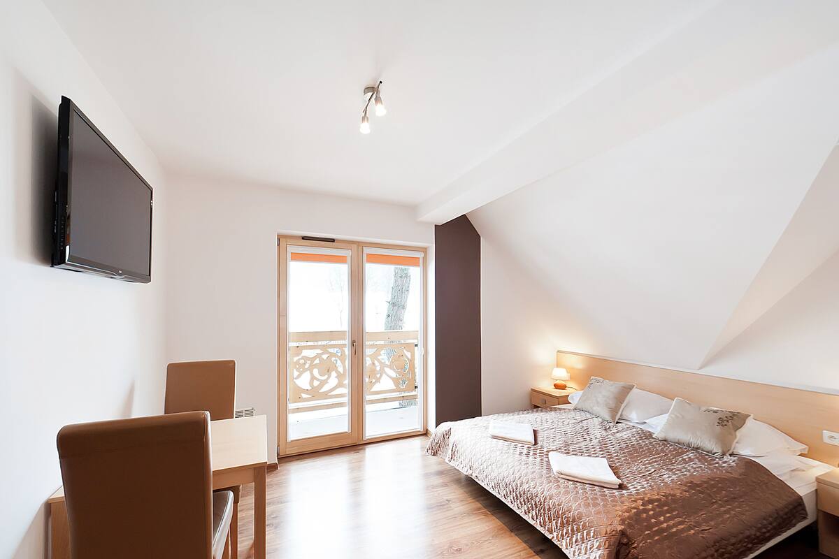 Een slaapkamer met een bed, een aan de muur bevestigde televisie, een bureau met een stoel en een raam met uitzicht op de omgeving.