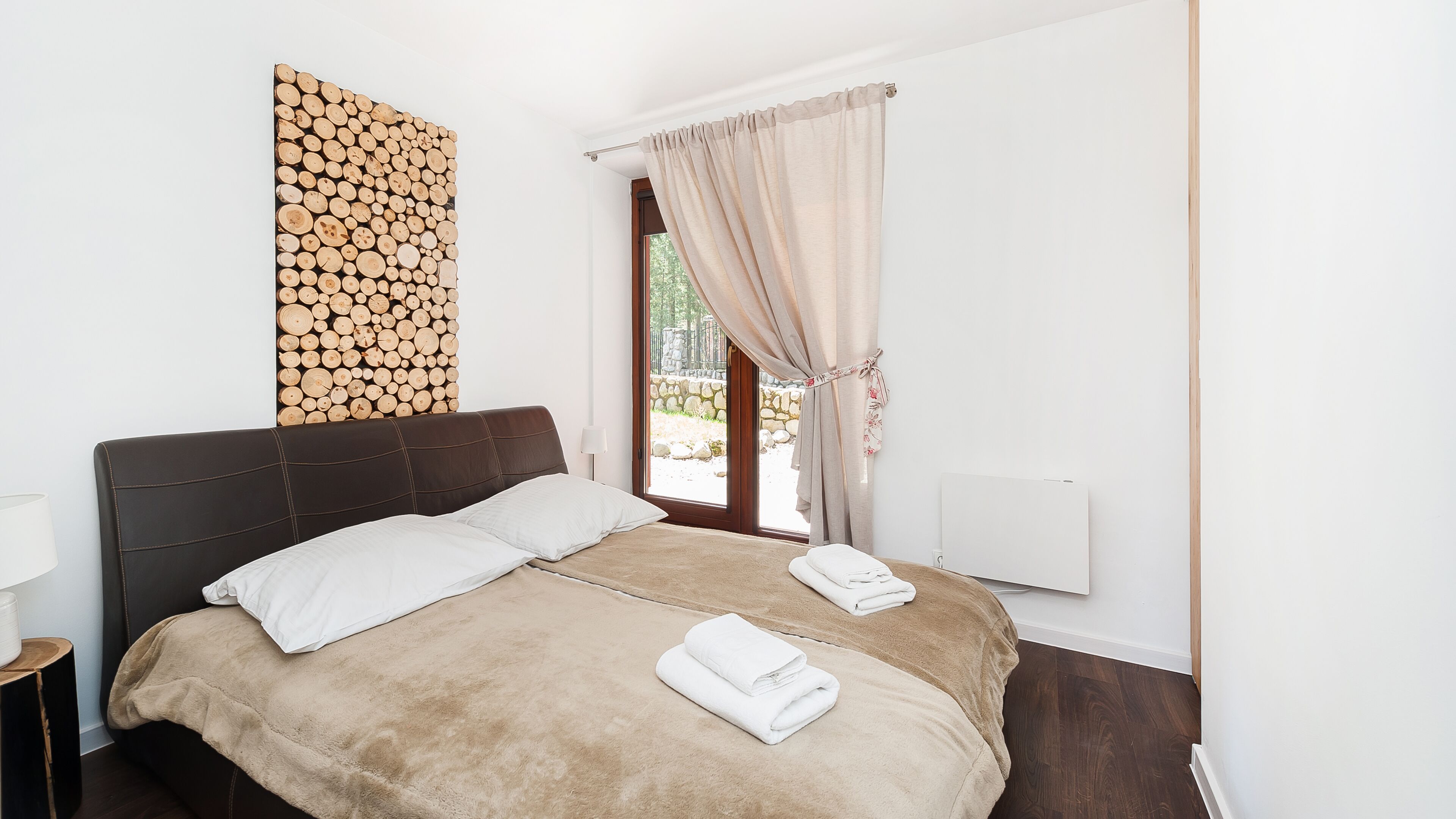 A bedroom with a large bed, a headboard, a wall-mounted log display, a window with curtains, and a bedside table with a lamp.