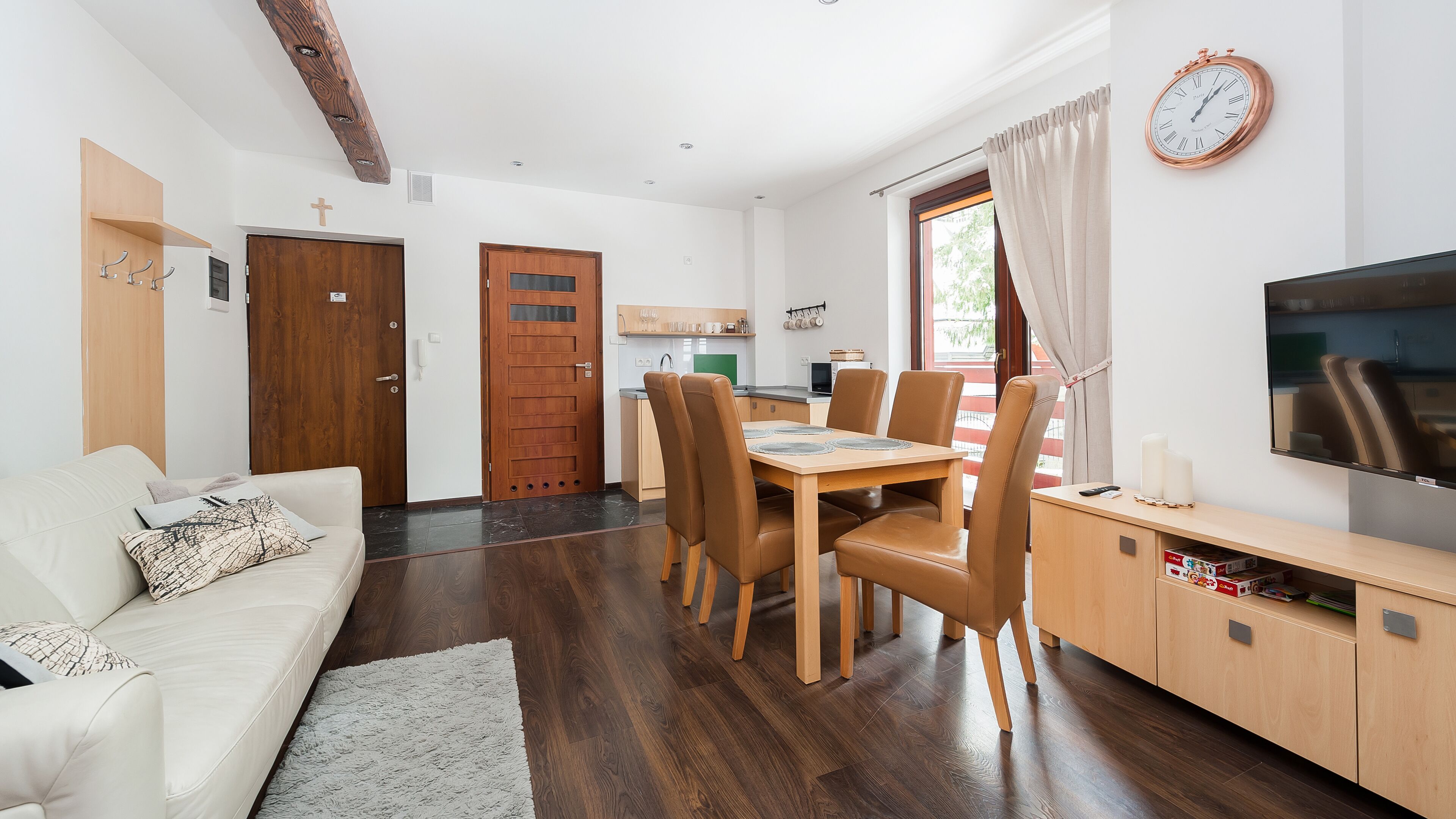 En moderne stue med sofa, spisebord med stole, fjernsyn og et køkkenområde med et vindue.