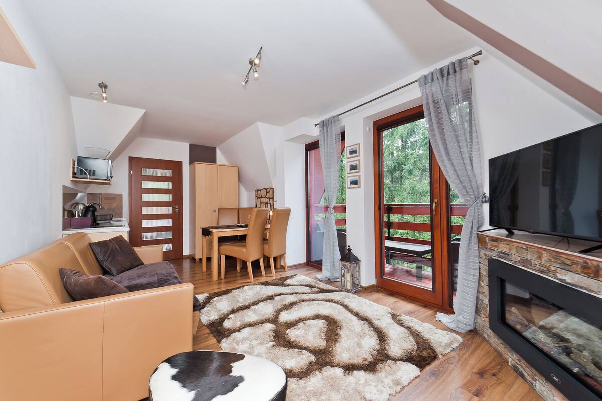 Een moderne woonkamer met een bank, televisie, open haard, eethoek en uitzicht op groen door de glazen deur.