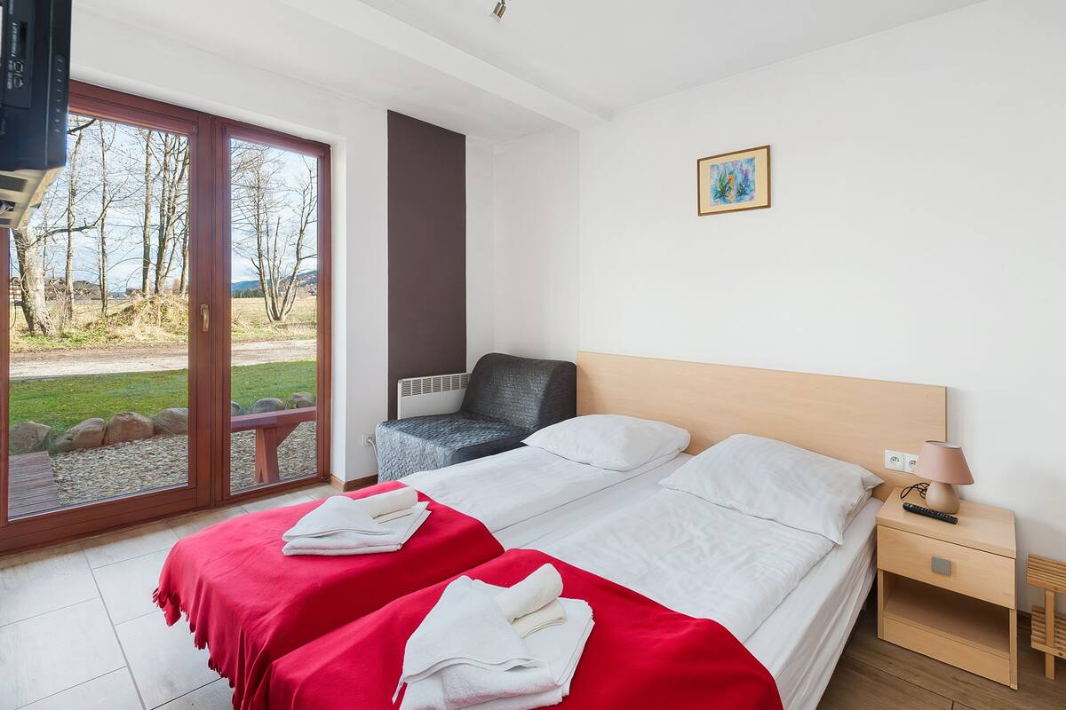 Een tweepersoonsbed met witte lakens, een rode plaid en een nachtkastje met een lamp.