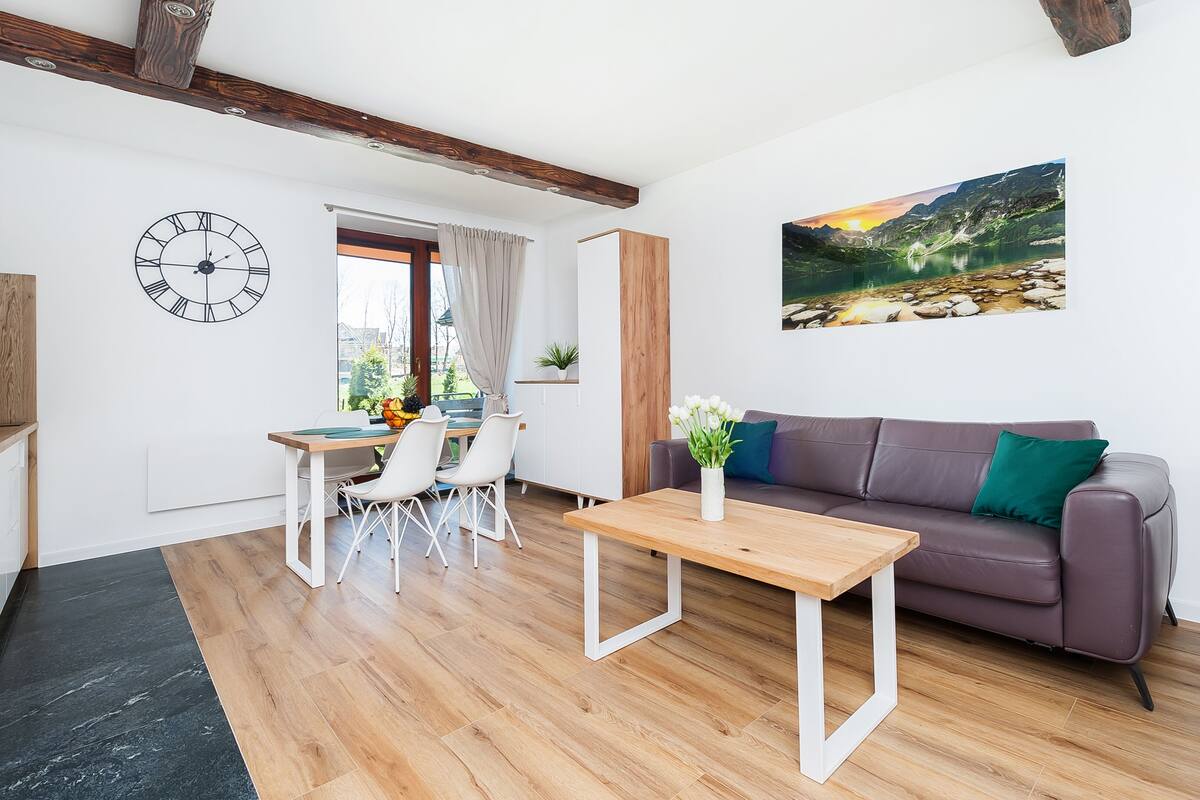 Een moderne woonkamer met een houten vloer, een eethoek met witte stoelen, een bank met kussens, een klok en een schilderij van een berglandschap.