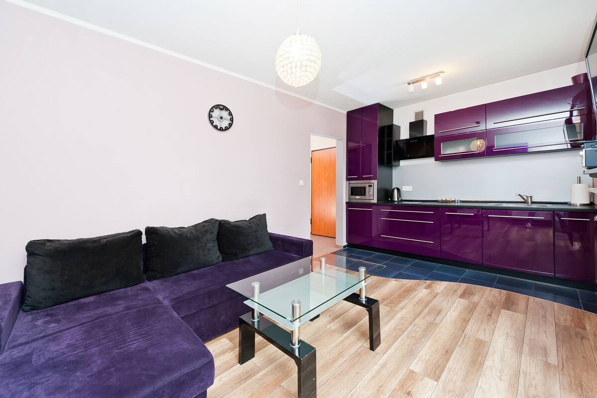 Een moderne woonkamer met een paarse bank, een glazen salontafel en een keuken met paarse kastjes.