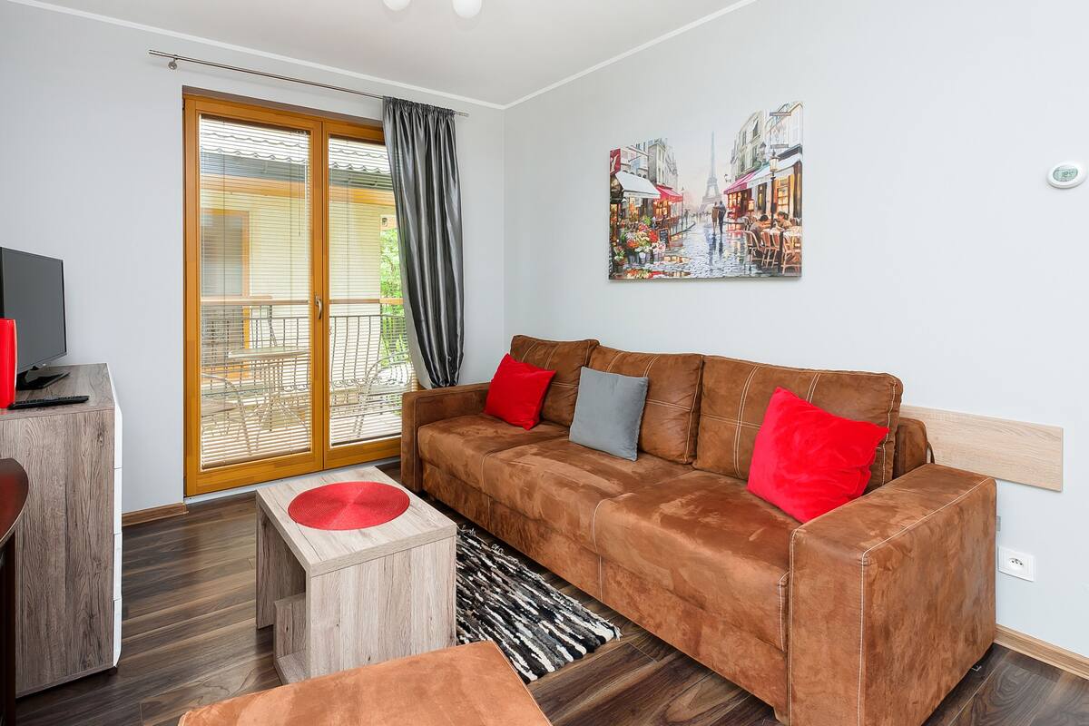 Een moderne woonkamer met een bruine bank, rode en grijze kussens, een houten salontafel, een tv op een houten dressoir en een schilderij van een straatbeeld.