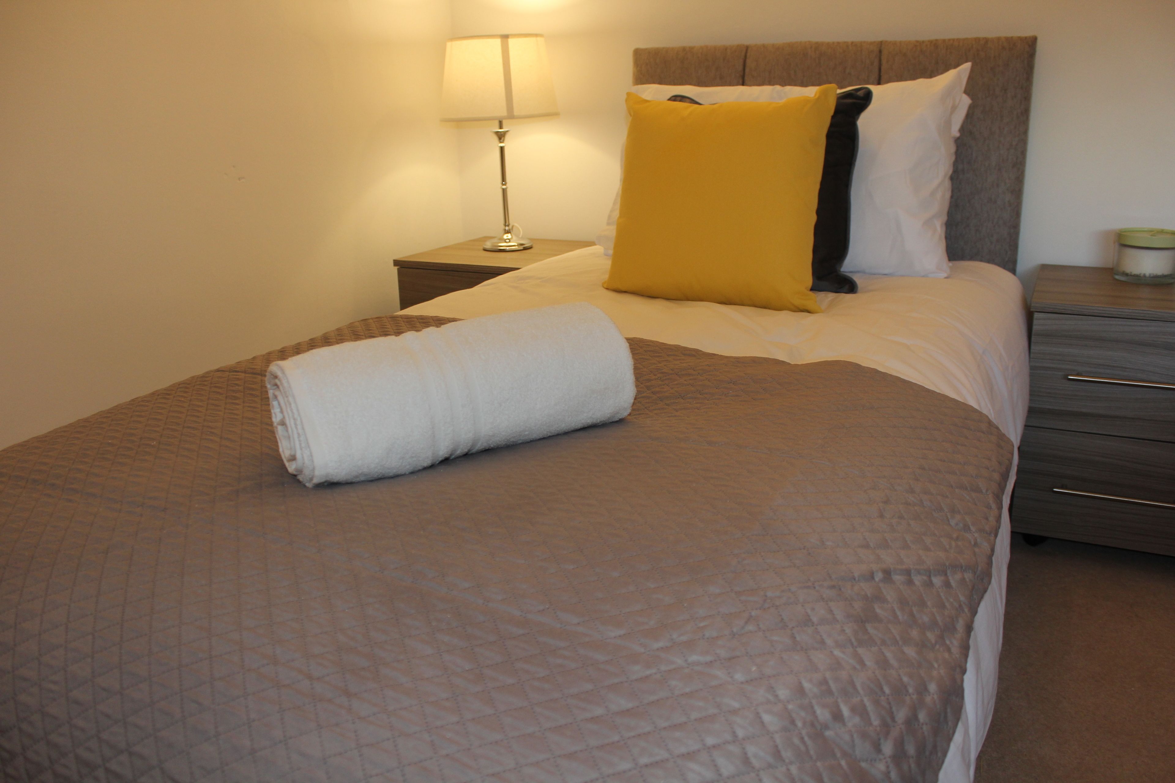 Una cama bien hecha con una toalla blanca enrollada, una lámpara de noche y una mesita de noche con una vela.